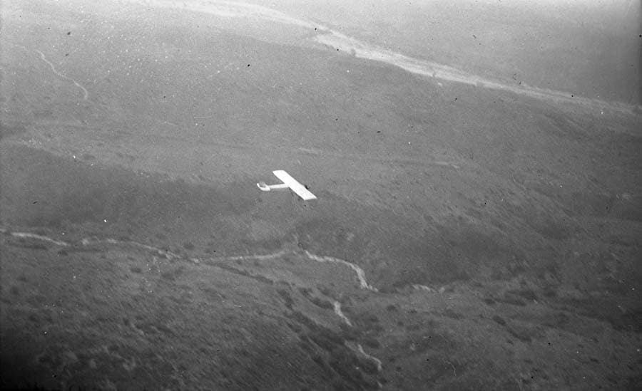 An air-to-air photograph taken from a chase plane showing a distant view of the fully completed Spirit of St. Louis flying over the California landscape during its departure.