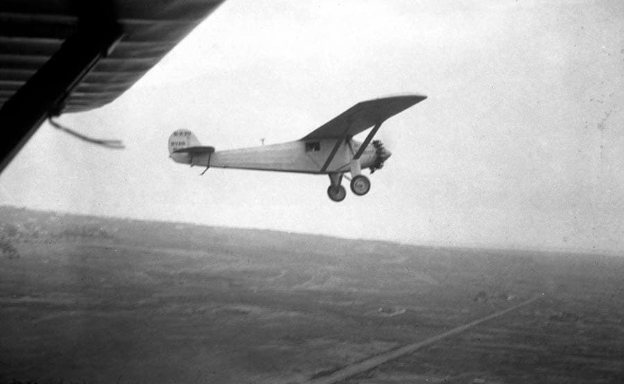 A close air-to-air photograph of the Spirit of St. Louis banking gently over the flat California terrain, taken from beneath the wing of the M-2 chase plane during its departure.