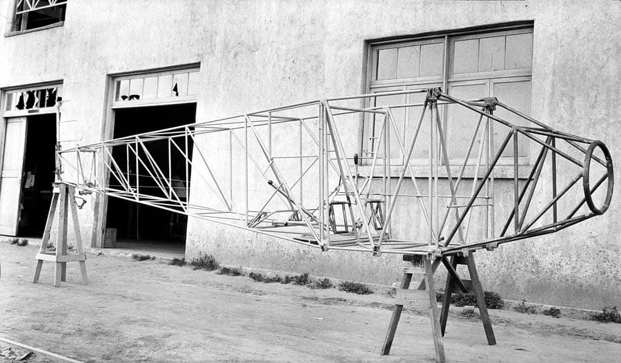 A full-length view of the bare tubular steel fuselage framework of the Spirit of St. Louis resting on sawhorses in front of the factory.