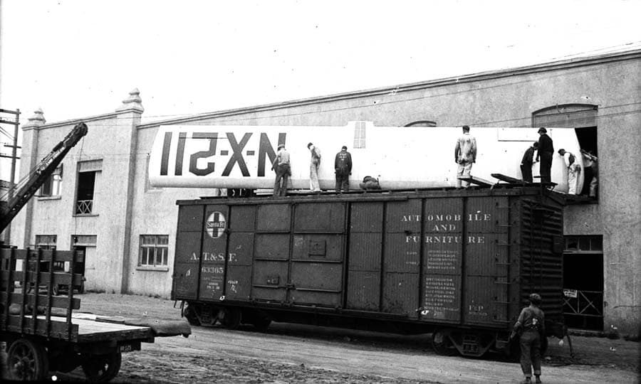 A crew of men carefully maneuvering the massive, completed Spirit of St. Louis wing out of a second-story factory window onto an unused train boxcar.