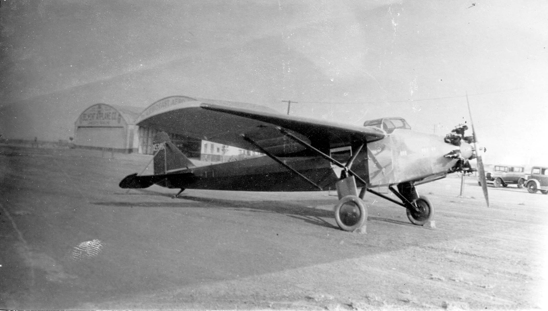 A three-quarter side profile of a Stinson SM-1 Detroiter monoplane with Wright Whirlwind engine parked outside airport hangars.