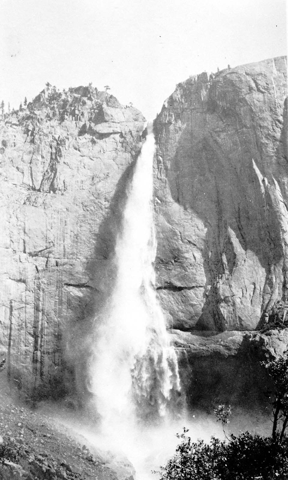 Upper Yosemite Fall plunging into its intermediate rocky basin with dense water vapor and mist drifting over evergreen trees.