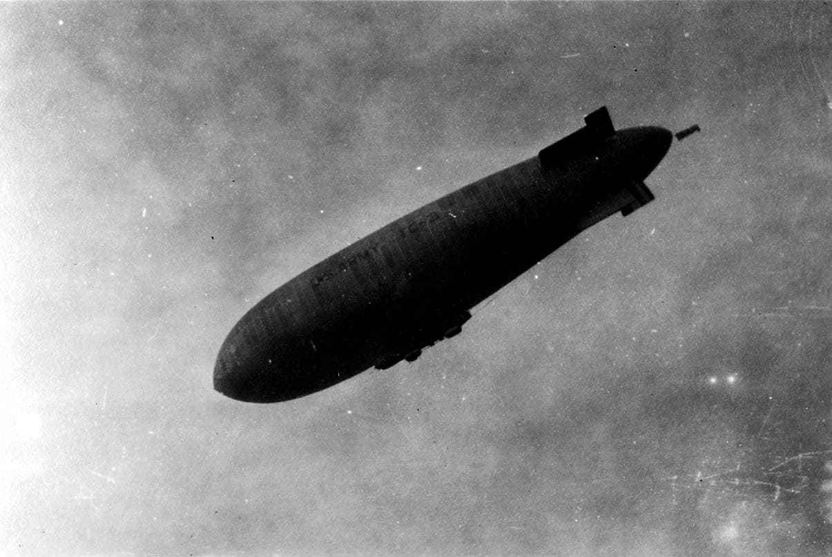 A dramatic low-angle telephoto view of the U.S. Army airship TC-2 showing the control gondola, empennage fins, and bold hull lettering.