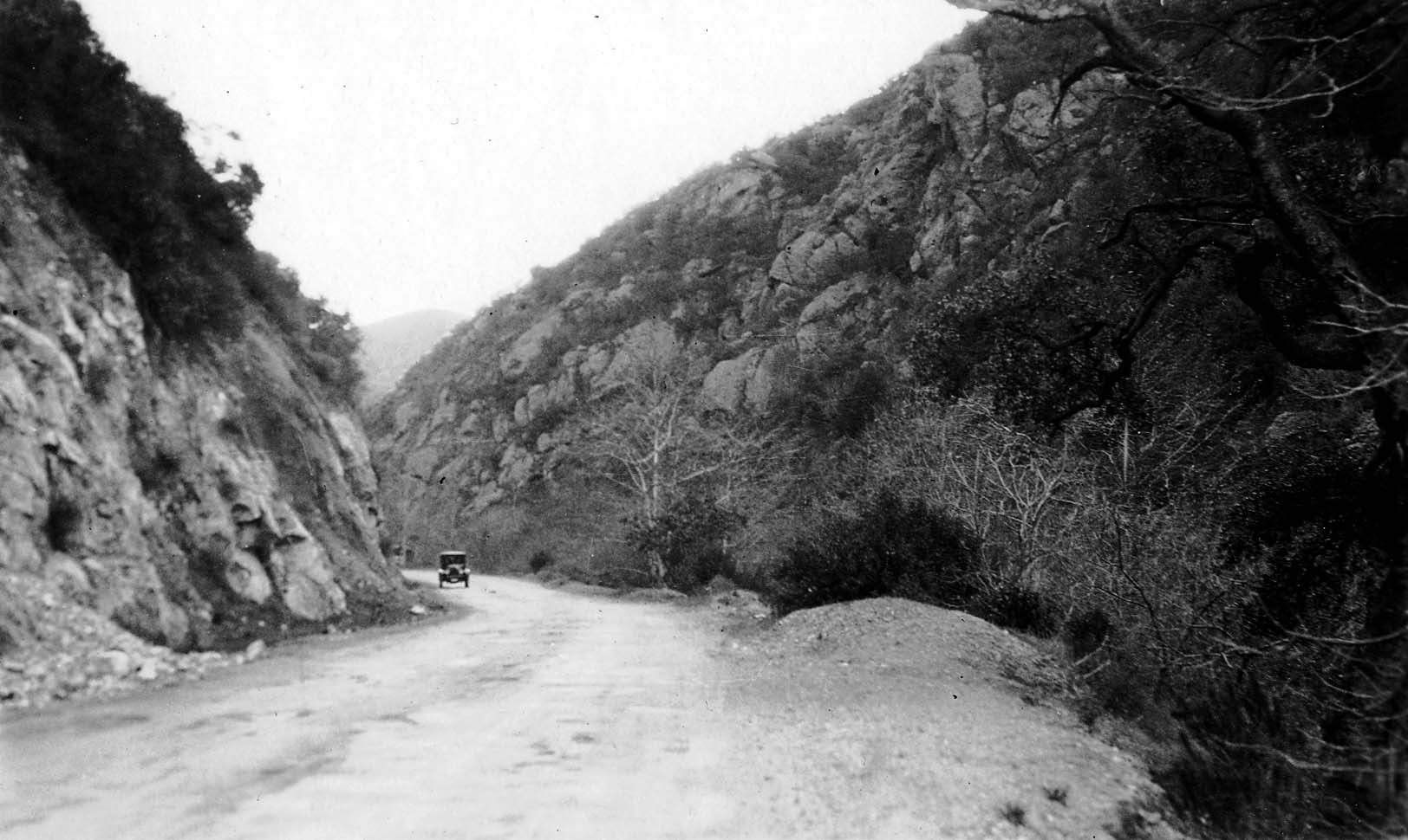 A lone vintage automobile driving along an unpaved road cut through a steep, rocky mountain gorge with rugged stone cliffs.