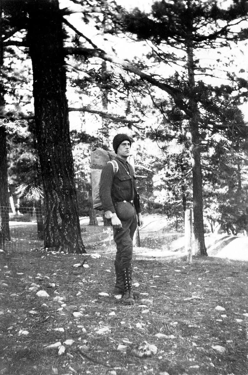 Donald A. Hall Sr. standing in profile among towering ponderosa pines wearing a heavy canvas pack, dark breeches, and a knit watch cap.