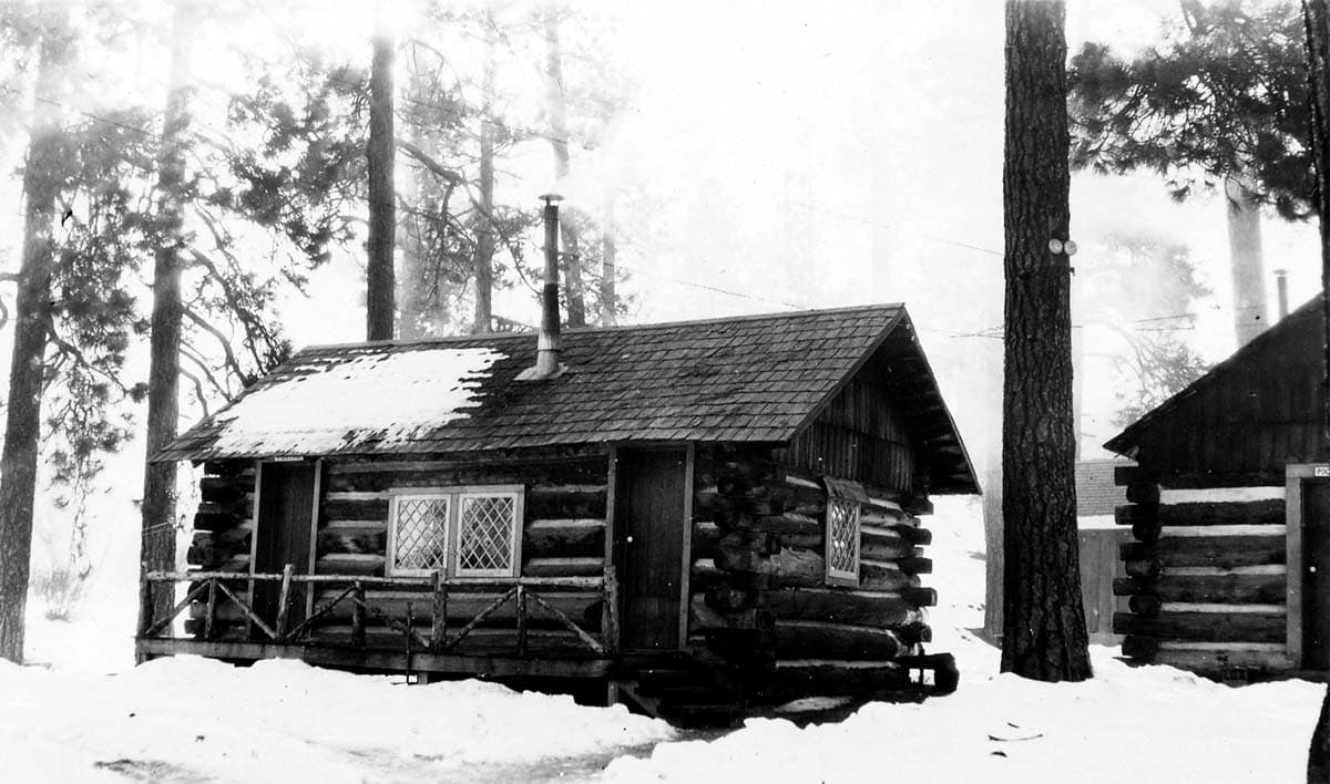 A rustic notched-timber log cabin with stone foundation and smoking chimney nestled in deep snow among pine trees.