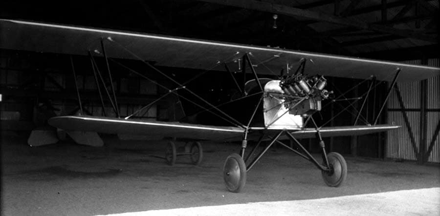 An open-cockpit biplane parked inside a dark corrugated metal hangar, showcasing an uncowled Wright-Hispano V-8 water-cooled engine.