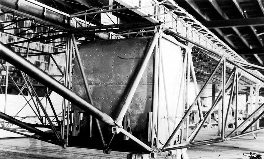 Factory view of uncovered fuselage frame, main fuel tank rivets, and wooden wing structure on sawhorses.