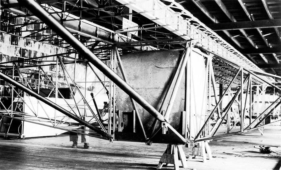 Factory view of fuselage frame and fuel tank showing shop worker standing behind framework.