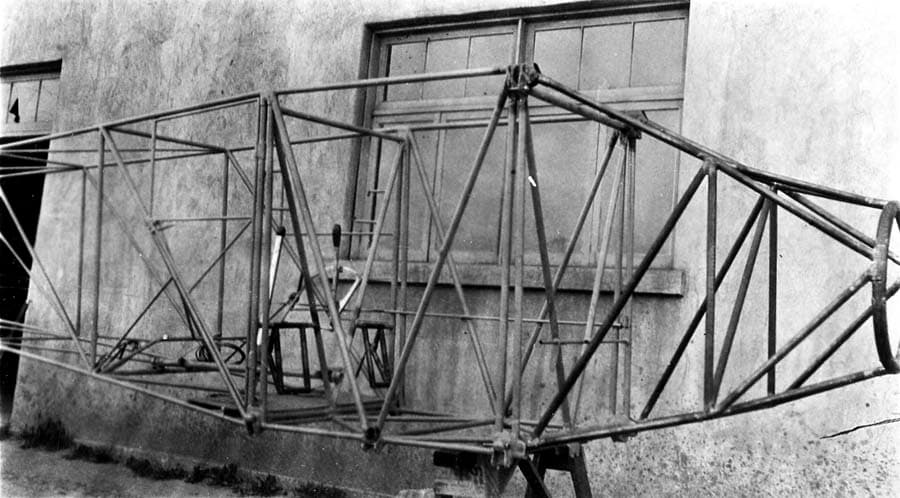 Uncovered welded steel-tube fuselage frame resting on sawhorses outside factory wall showing cockpit and fuel tank bay.