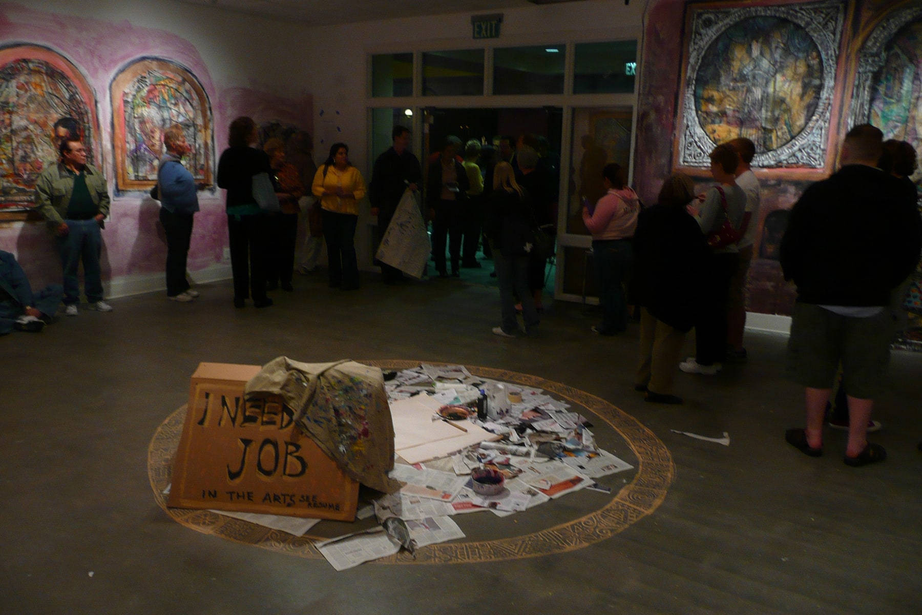 Visitors gather in a gallery around colorful paintings and a circular installation of newspapers and art materials.