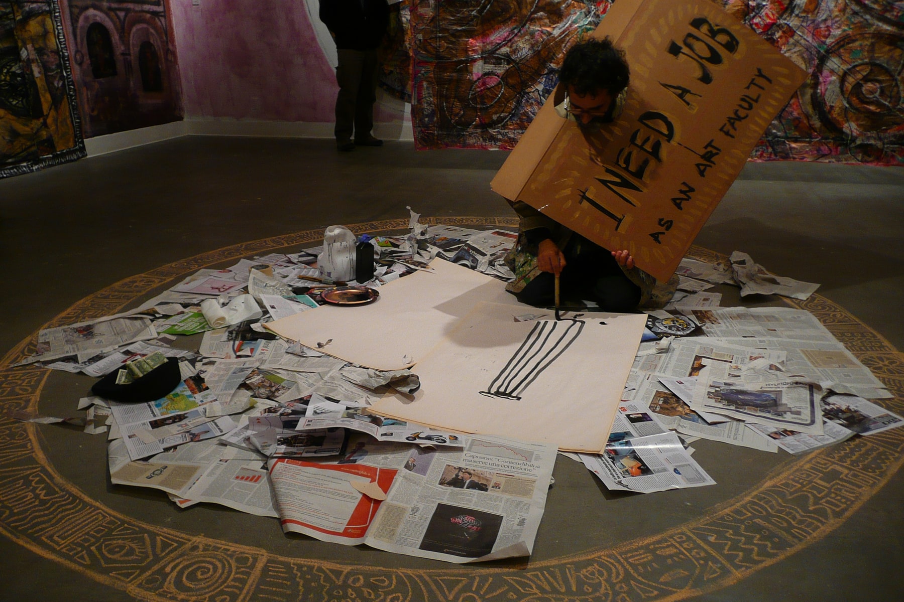 A performer kneels on newspapers to draw a column in black ink, wearing a cardboard sign that reads I need a job as an art faculty.