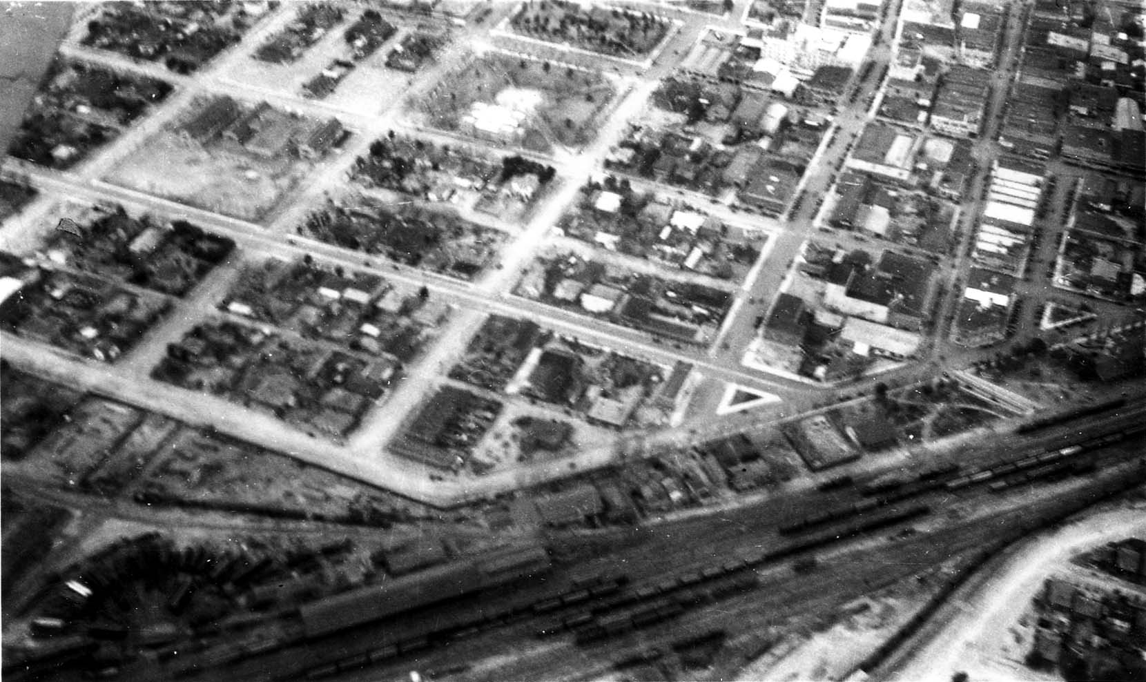 An aerial photograph looking down at a gridded city street layout with a large railyard and train cars in the foreground.