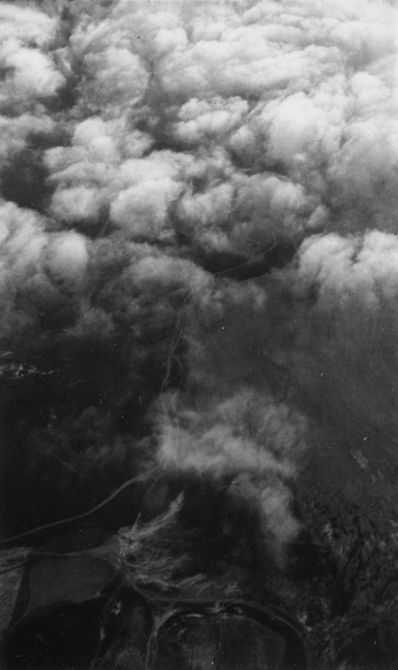 An aerial view from an aircraft cockpit looking down through billowing cumulus cloud banks to the terrain and winding roads below.