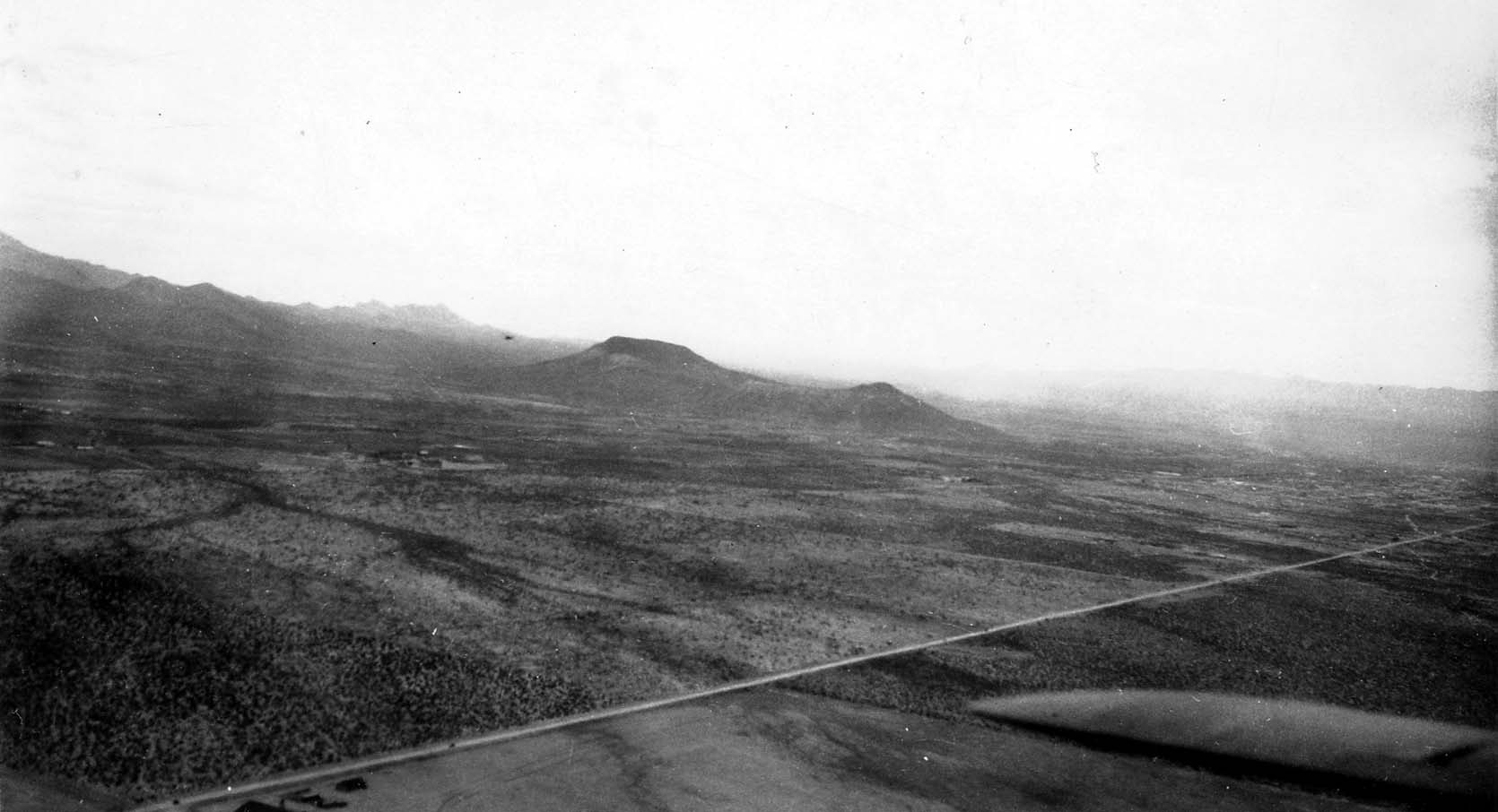 An aerial view looking past the edge of an aircraft wing over flat desert scrubland toward a distant butte or hill.