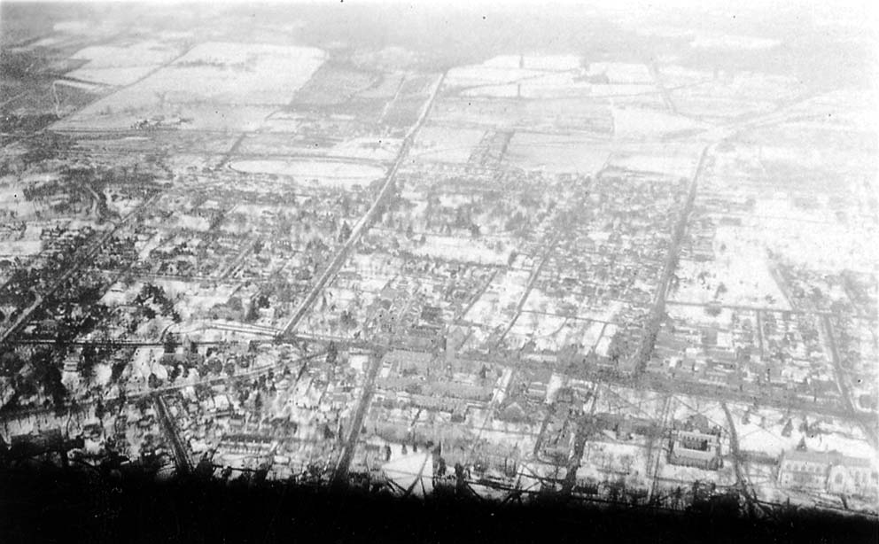 A high-altitude aerial photograph looking straight down at a sprawling, snow-covered town with distinct roads and housing blocks.
