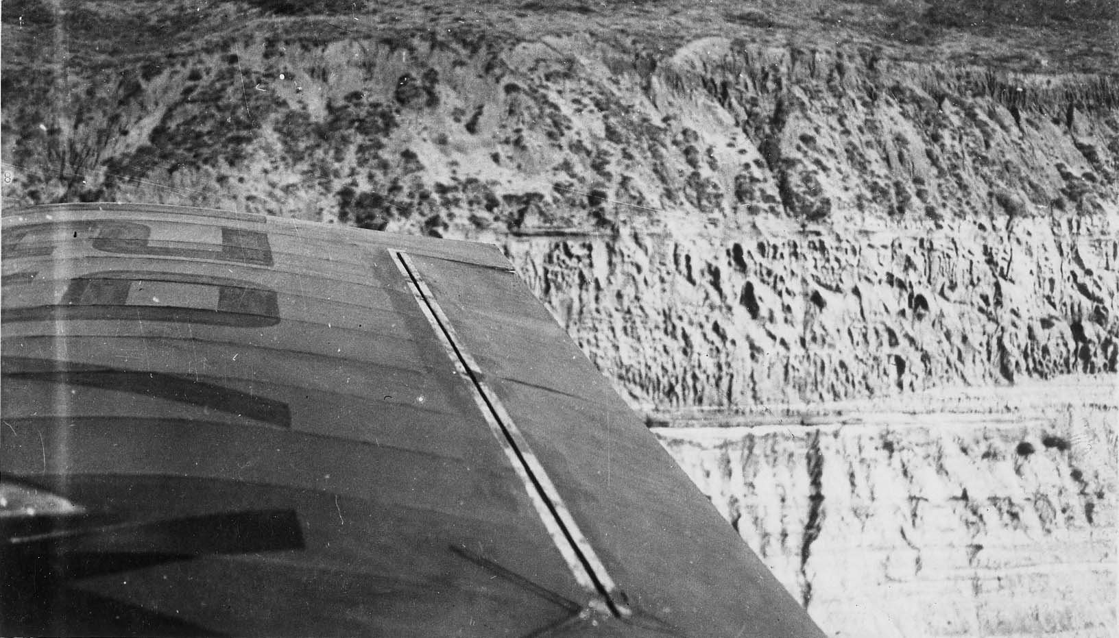 An aerial view from an aircraft cockpit looking past the wing and aileron directly down at the sheer, eroded coastal sandstone bluffs.