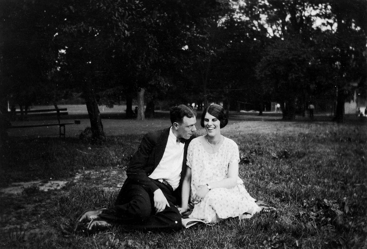 Alice Hall in a floral summer dress laughing beside a smiling companion in a suit and bow tie seated on a grassy park lawn under shady trees.