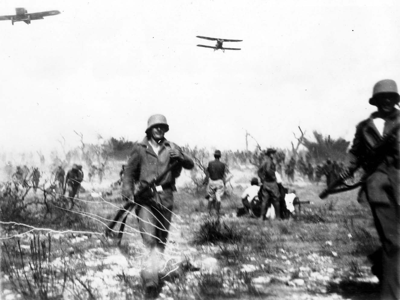 Doughboy soldiers charging through brush terrain as a single-engine military biplane buzzes at treetop level during tactical war games.