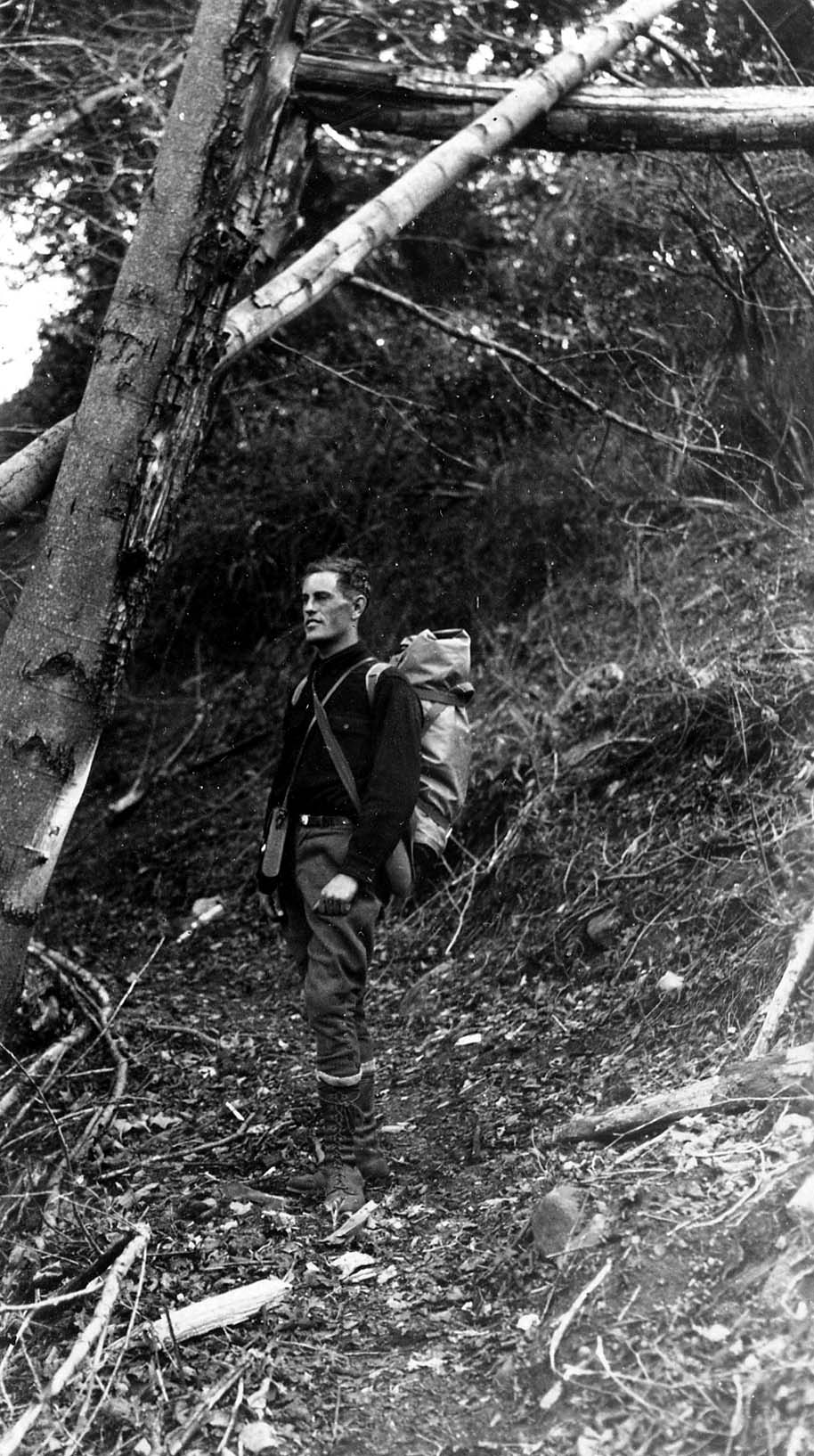 A full-length self-portrait of Donald A. Hall Sr. on a wooded mountain trail carrying a heavy canvas external-frame backpack and bedroll.