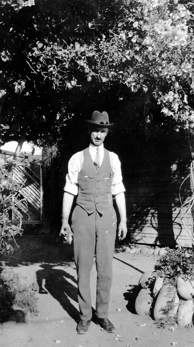 Betty Walker's father dressed in a tailored vest, tie, and fedora holding a camera under a shaded garden arbor on a wedding day.