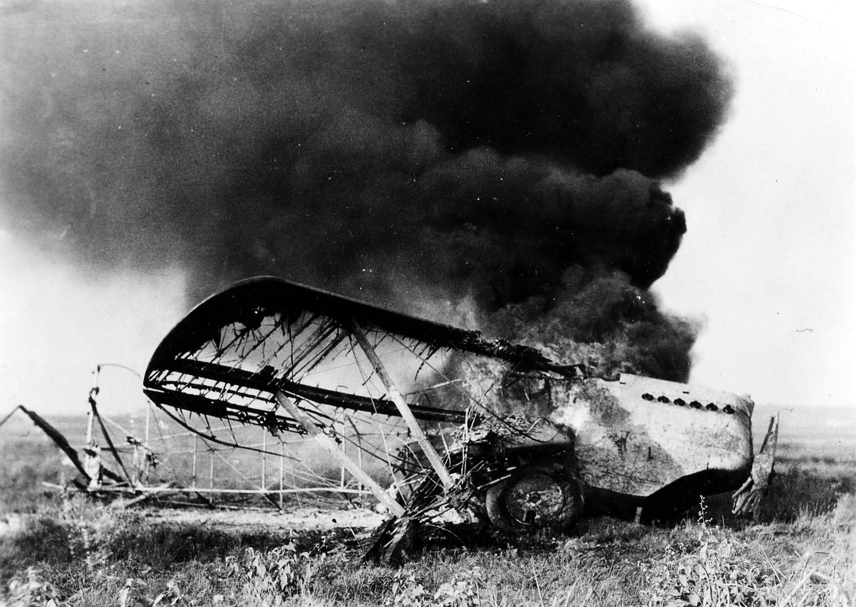 A crashed biplane engulfed in intense flames with thick black smoke billowing into the sky, showing burned fabric ribs and exposed V-engine.