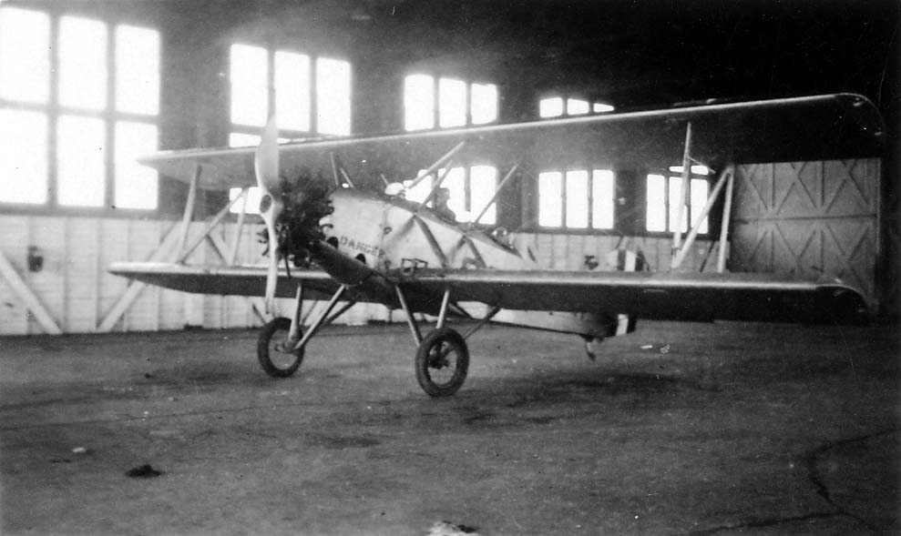 An open-cockpit biplane inside a brightly lit hangar with the word DANGER stenciled in bold block letters near the propeller arc.