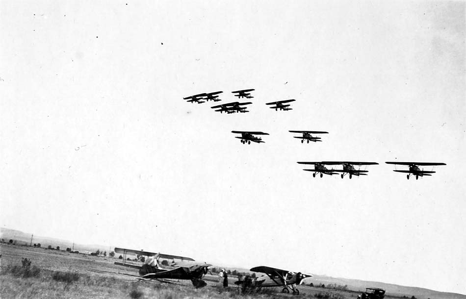A formation of vintage biplanes flying low over a dirt airfield, with the experimental Hall X-1 low-wing monoplane parked in the foreground.