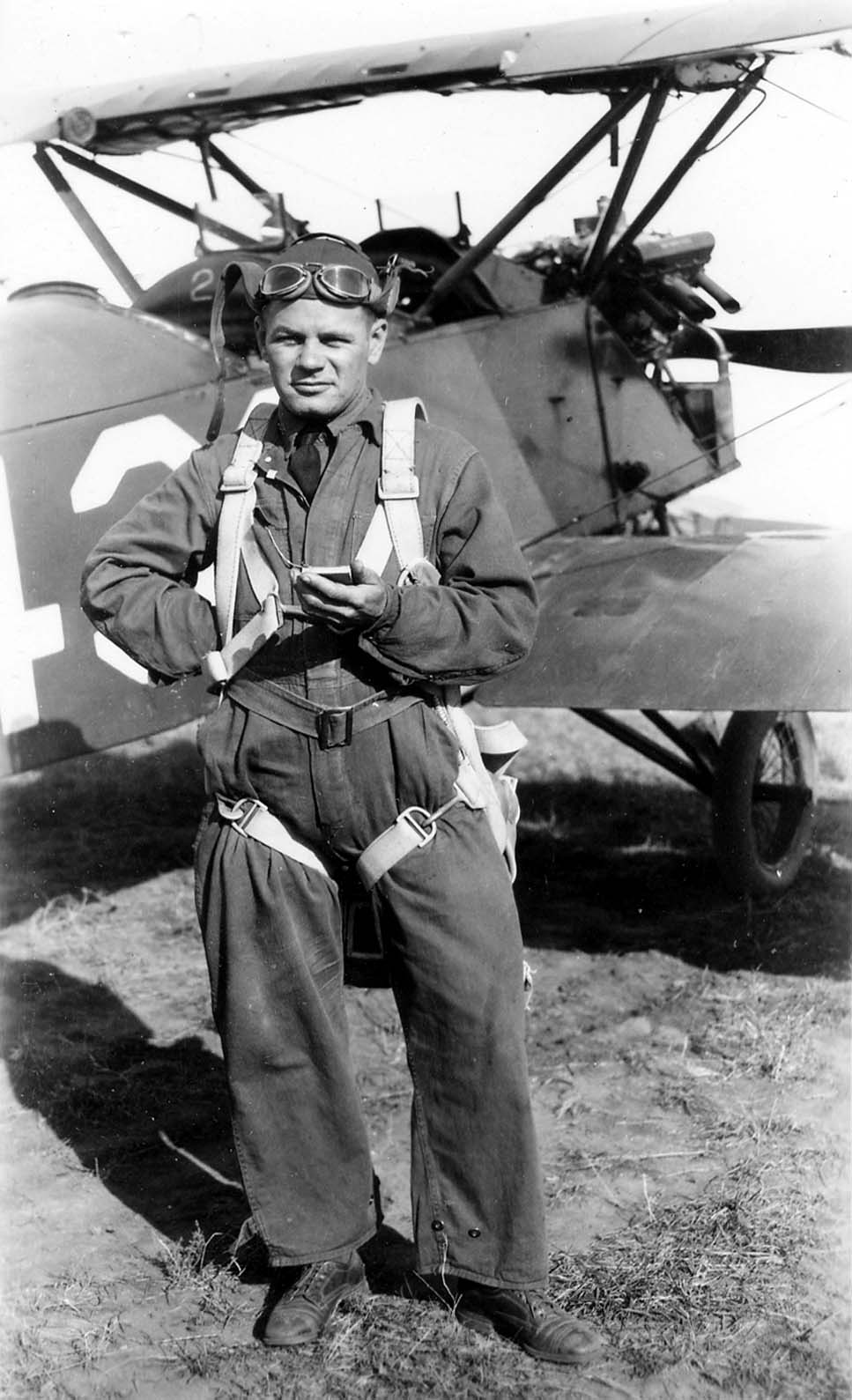 A vintage military aviator in flight suit, helmet, and parachute harness standing on an airfield in front of an open-cockpit biplane.