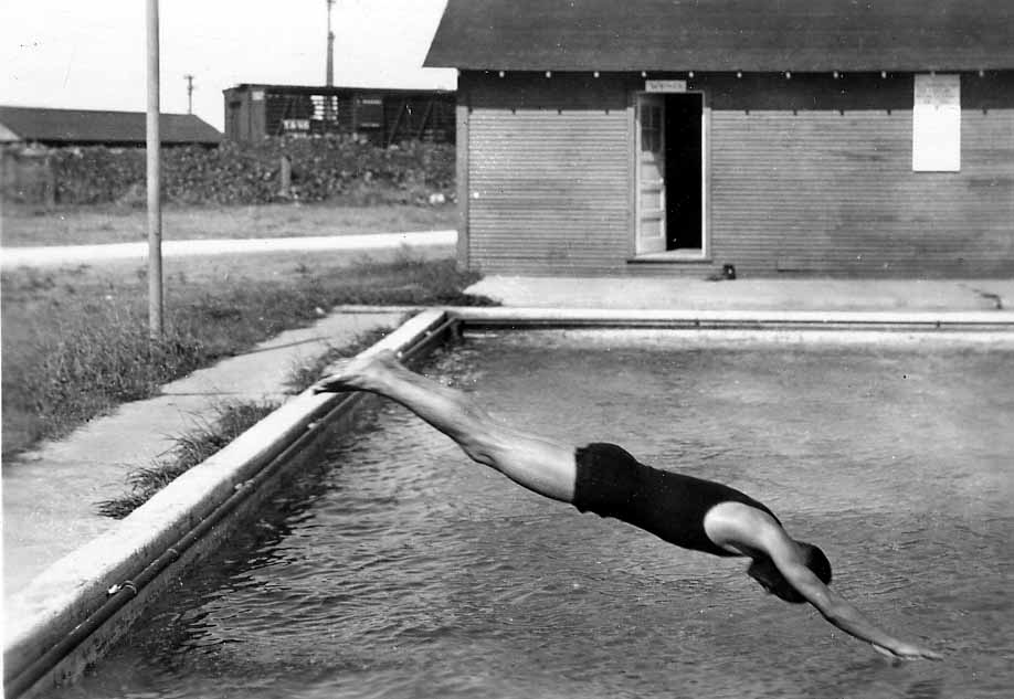 A diver fully extended in a shallow, head-first dive just inches above the water's surface, with a wooden bathhouse in the background.