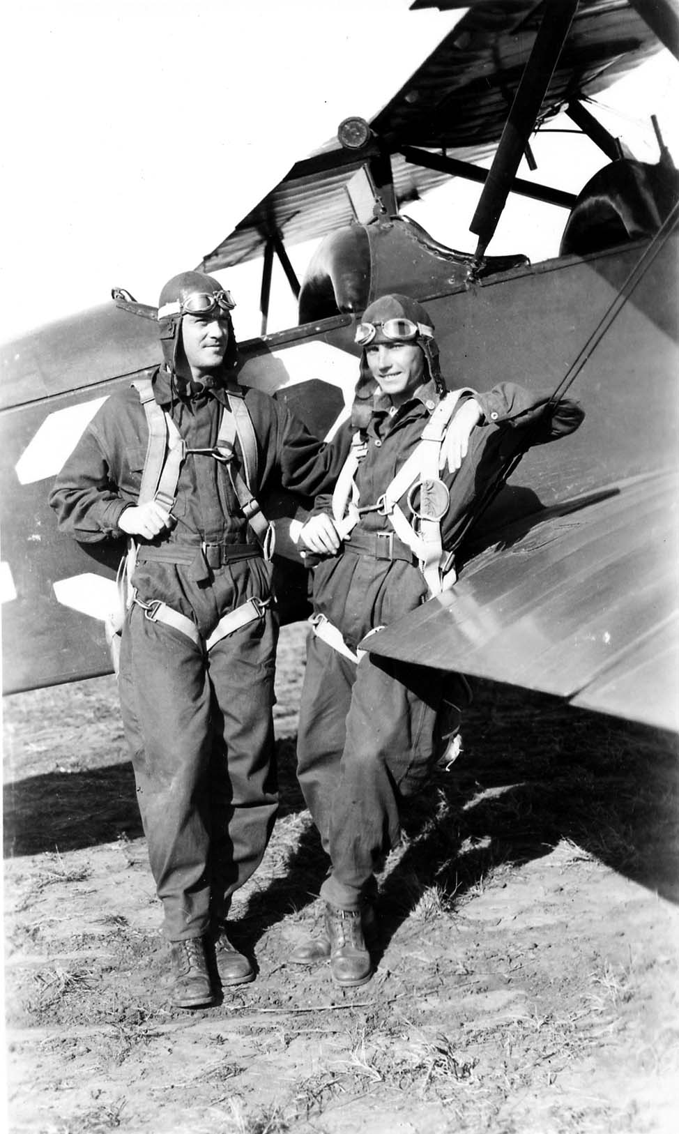 Two flight cadets wearing flight suits, leather helmets, and parachute harnesses, leaning casually against the wing of a biplane.