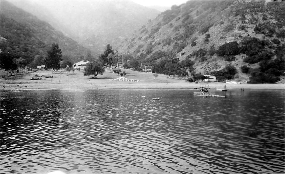 A view across the calm waters of a mountain lake toward Camp Whittier, featuring camp buildings and signage along the shoreline beneath steep hills.
