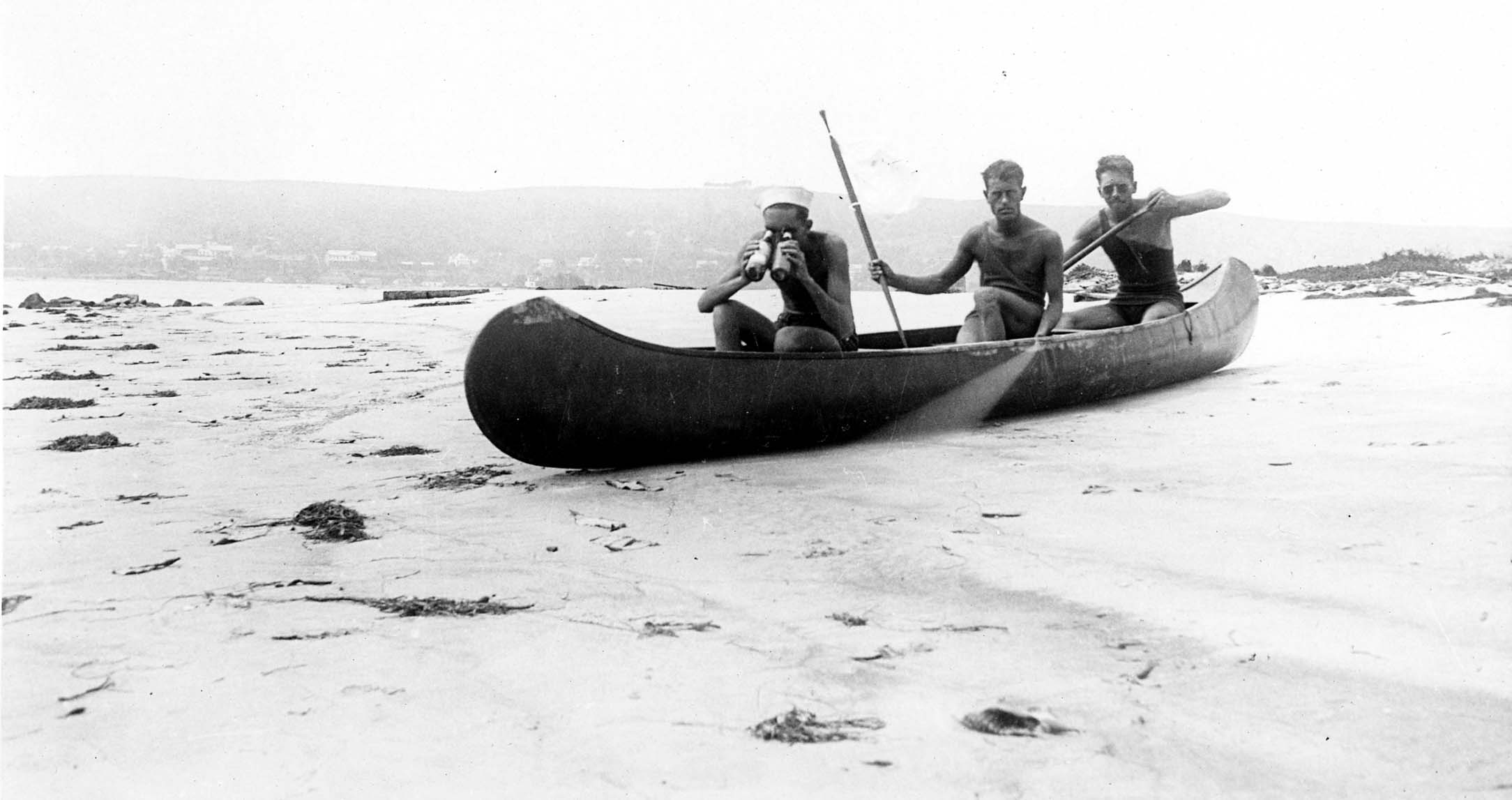 Three men sitting in a canoe on a sandy beach, with one looking through binoculars and a camera trigger string visible.