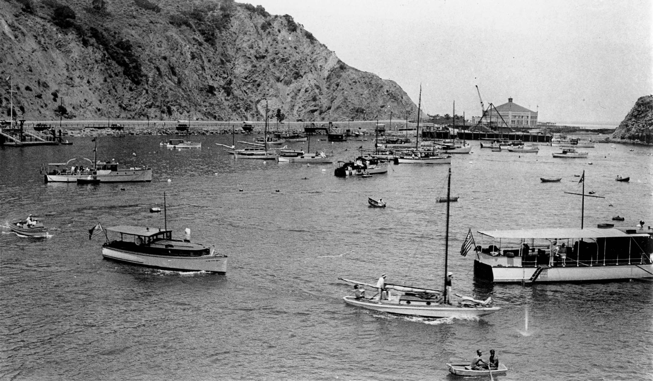 A high-angle view looking out over a bustling harbor filled with sailboats, rowboats, and small ferries, with a large, ornate building on the far shore.