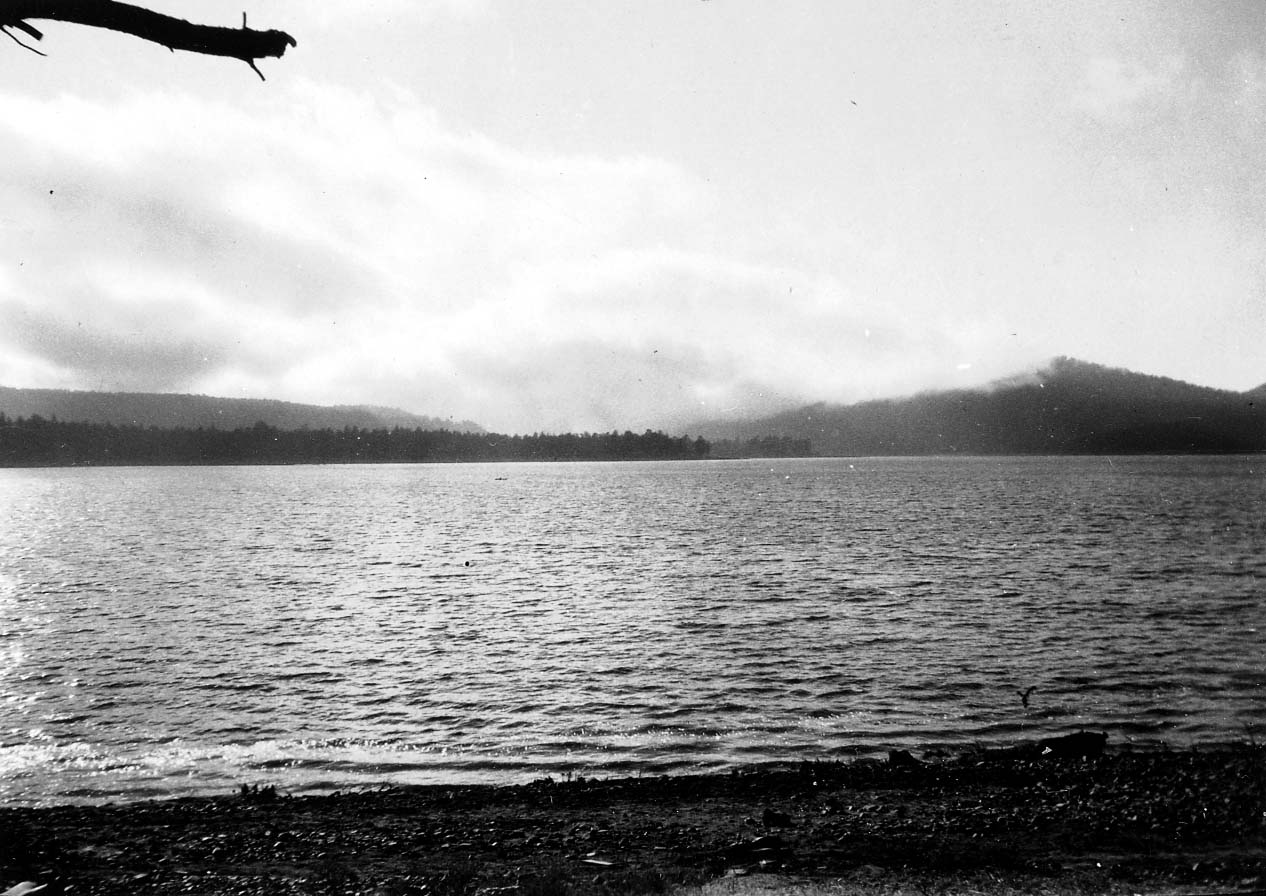 A wide view of choppy lake water along a pebbled shoreline under low-hanging morning clouds blanketing the distant mountain ridges.
