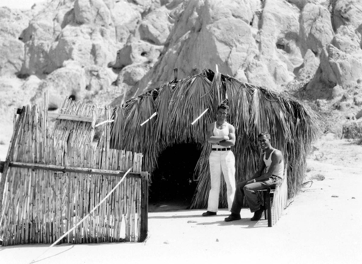 Two men in tank tops posing in front of a rustic palm-frond hut in a rocky desert landscape.
