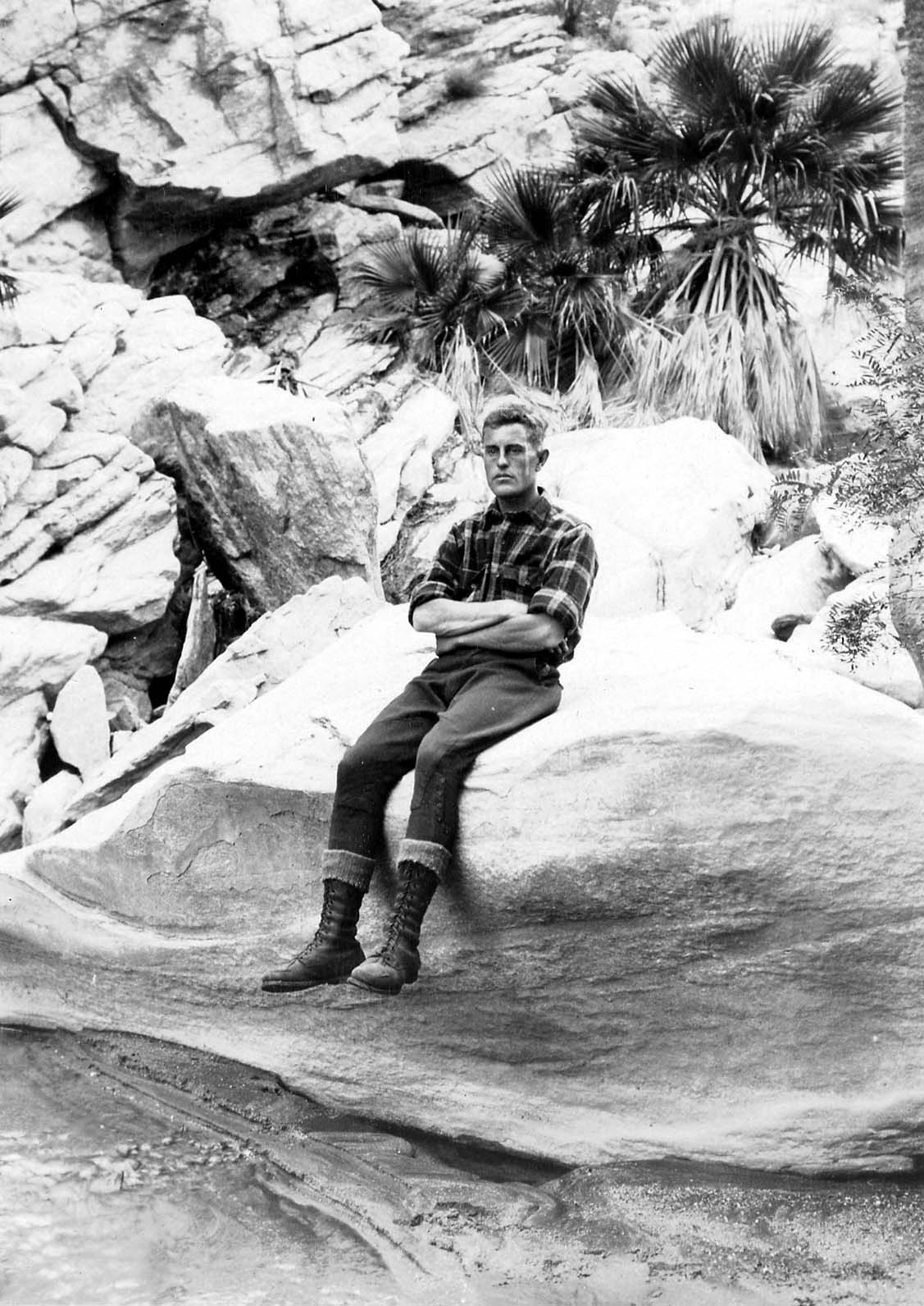 Donald A. Hall Sr. wearing a plaid shirt and tall boots, sitting with arms crossed on a white boulder near desert palm trees.