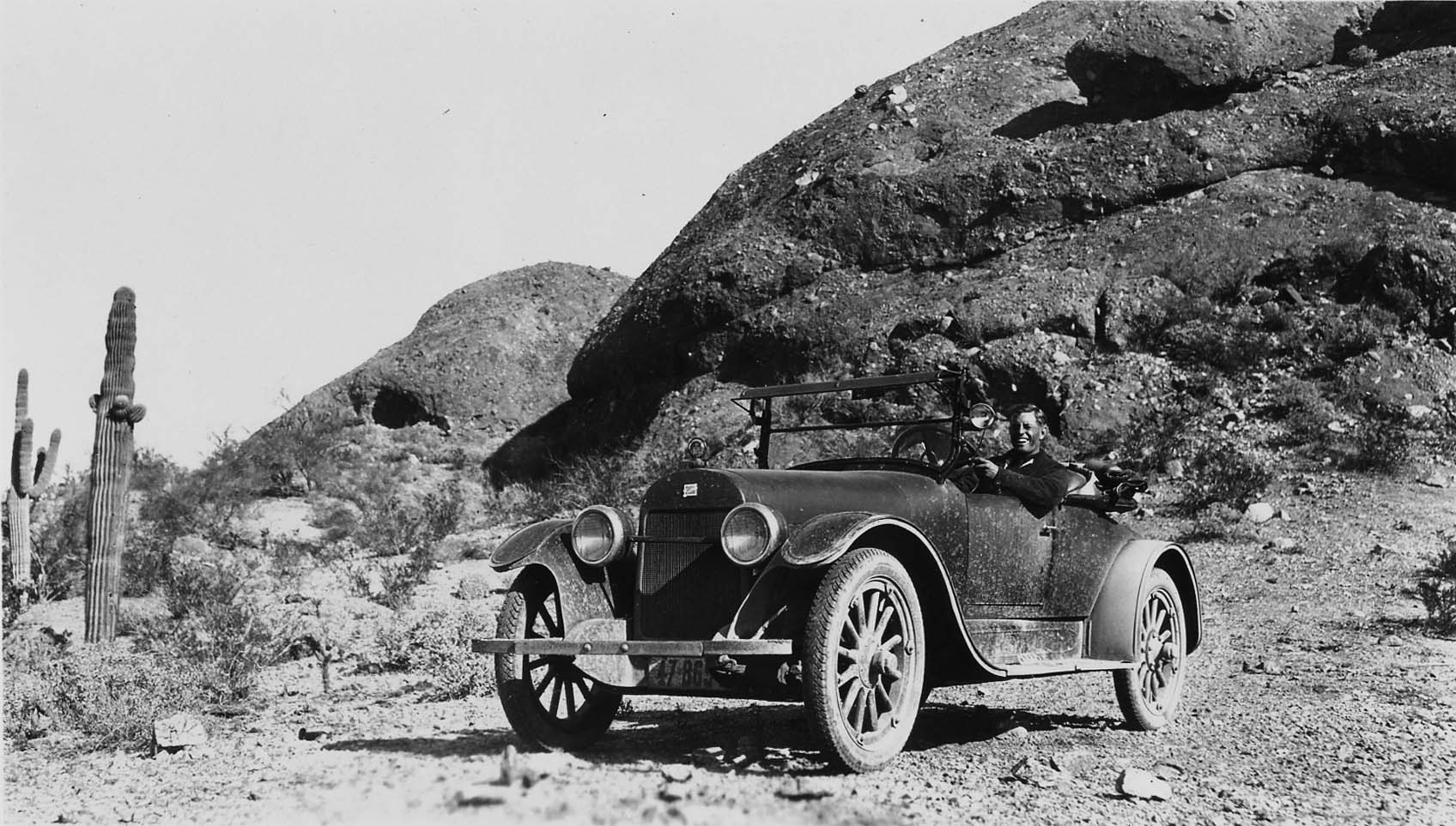 Donald A. Hall Sr. driving a vintage car on a desert dirt road past a large saguaro cactus and rocky hills.