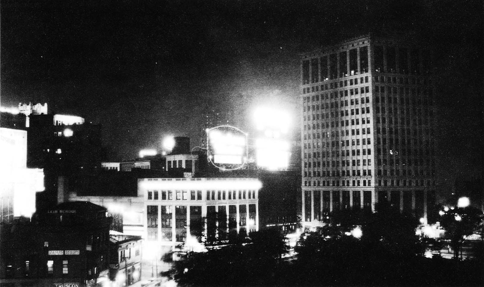 A night photograph looking across glowing illuminated commercial storefronts toward the towering David Whitney Building rising into the dark Detroit sky.