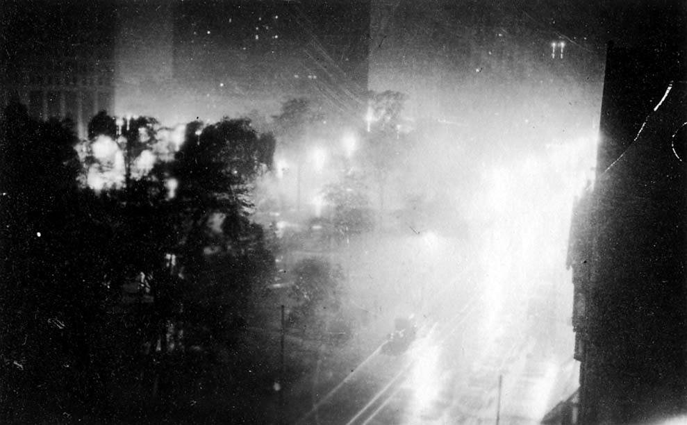 A high-angle nighttime view looking down through mist and fog over illuminated streets, glowing lampposts, and park trees in Downtown Detroit.