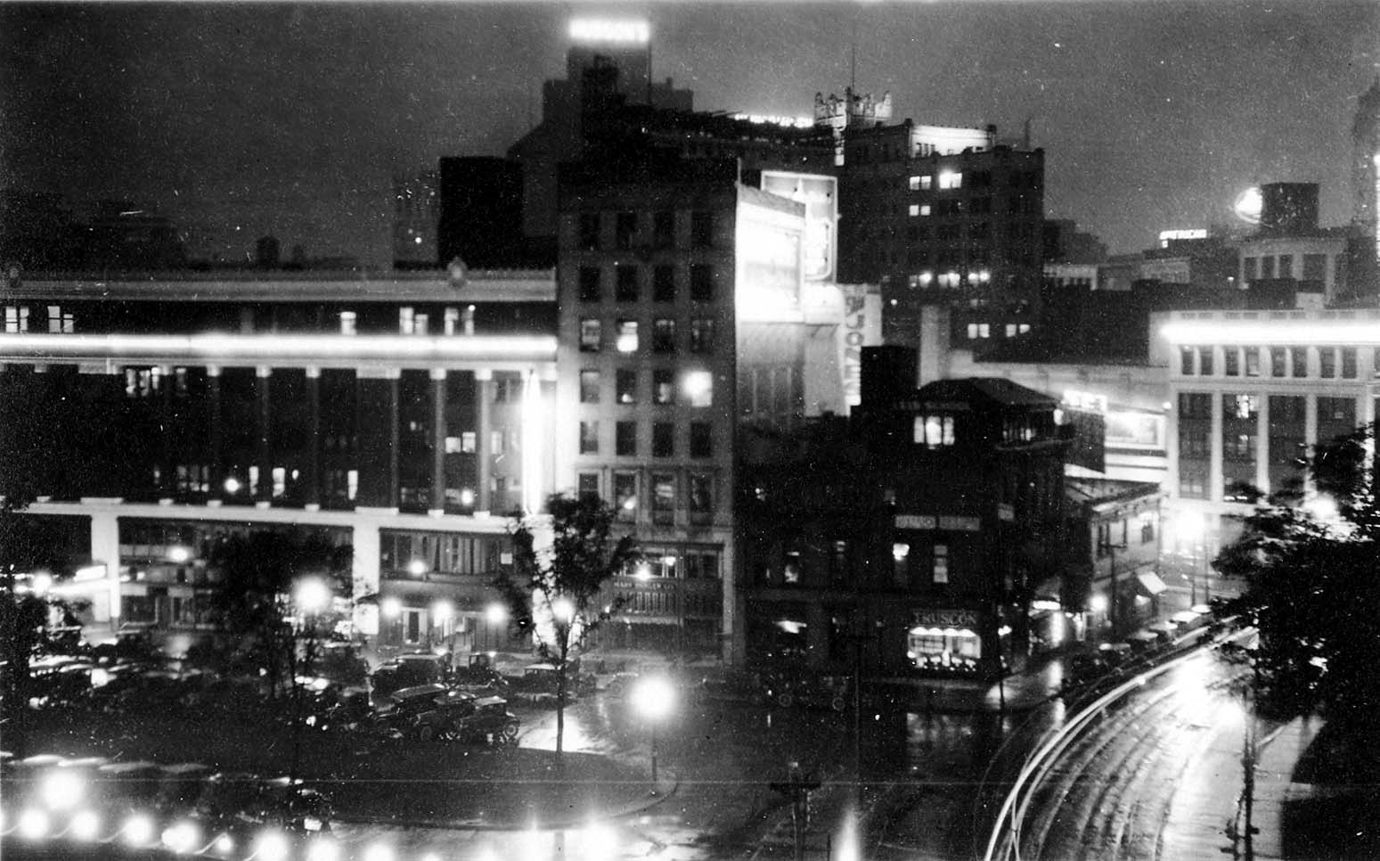 An elevated night streetscape showing parked vintage automobiles lining Grand Circus Park, glowing storefronts for Truscon, and the distant illuminated rooftop sign for Hudson's.