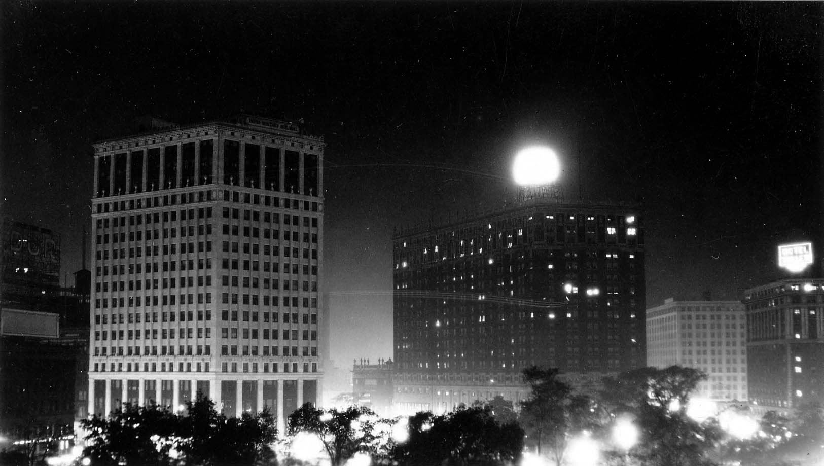 An expansive night skyline view overlooking the dark canopy of Grand Circus Park toward the brightly illuminated David Whitney Building and the Hotel Statler with its glowing rooftop globe.