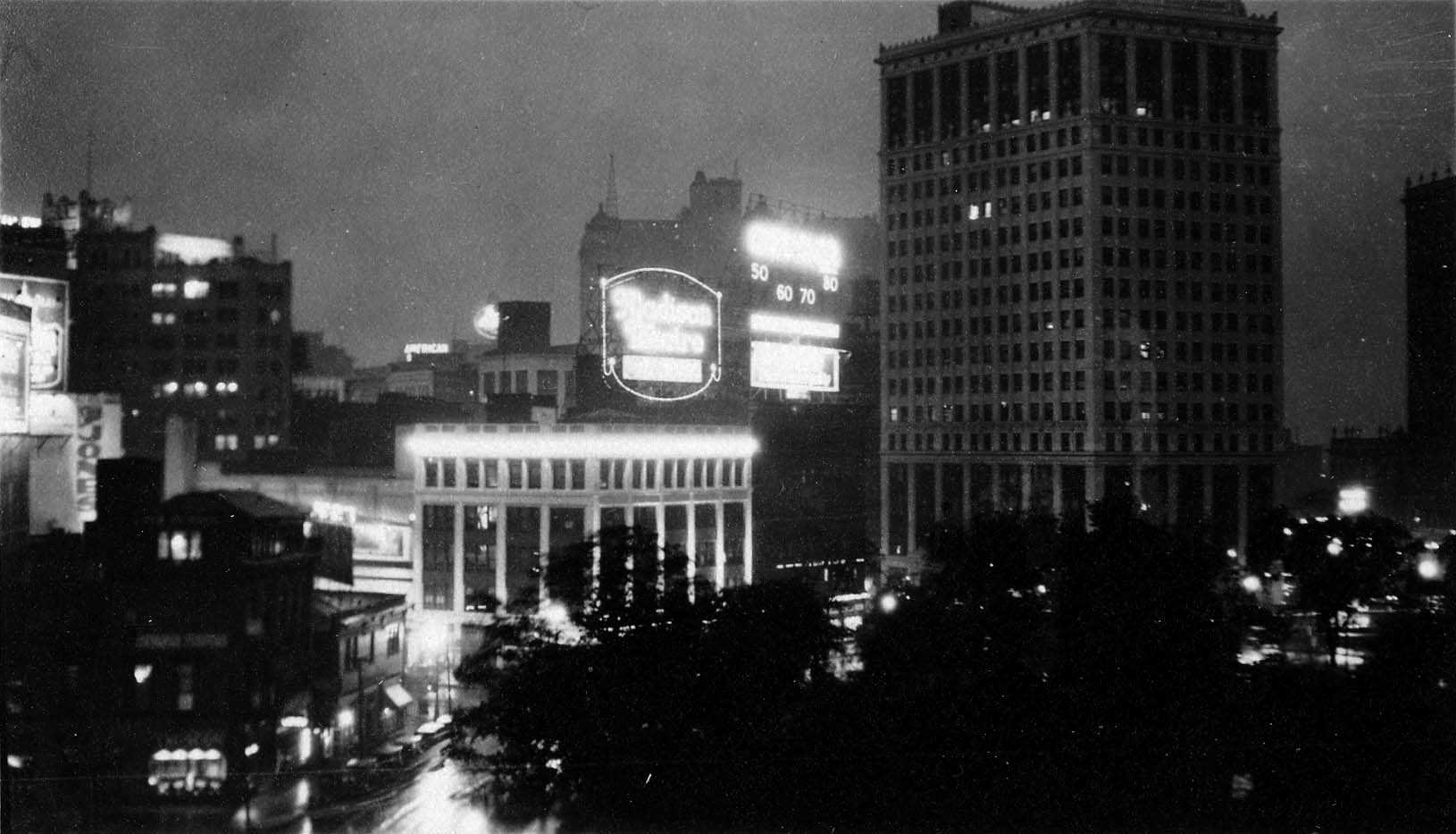An elevated view across Grand Circus Park at night, showcasing the glowing neon marquee of the Madison Theatre, illuminated billboards, and the towering David Whitney Building.