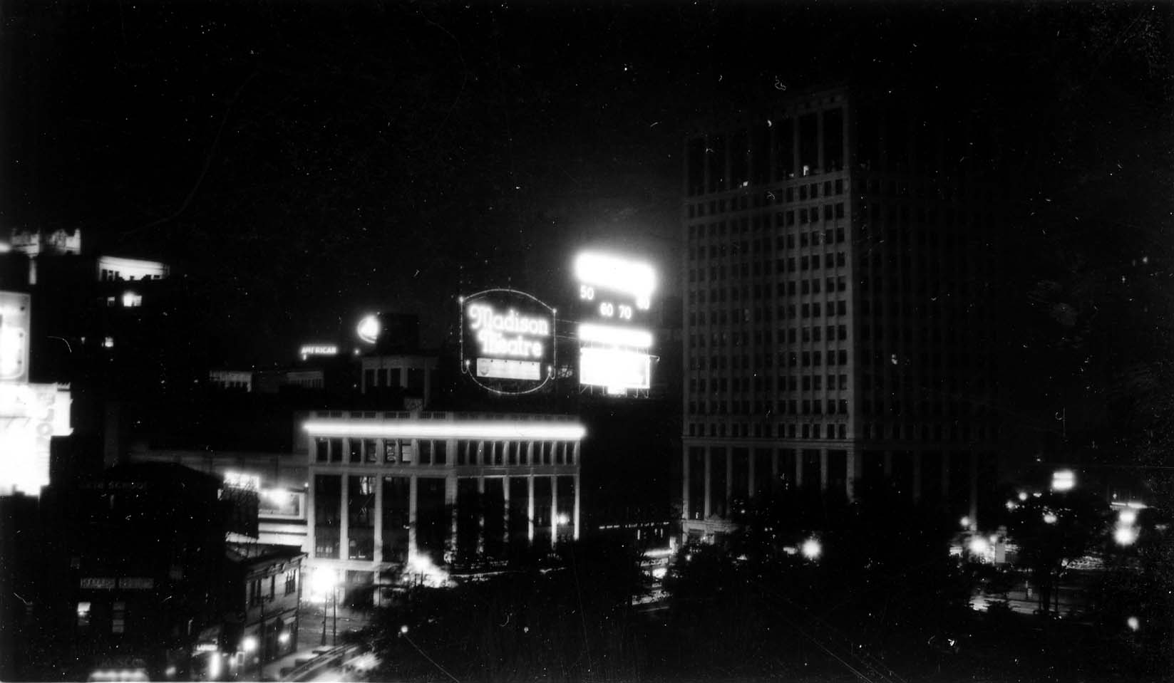 A high-contrast nighttime view centered on the brightly lit oval neon marquee of the Madison Theatre, with the dark silhouette of the David Whitney Building rising to the right.