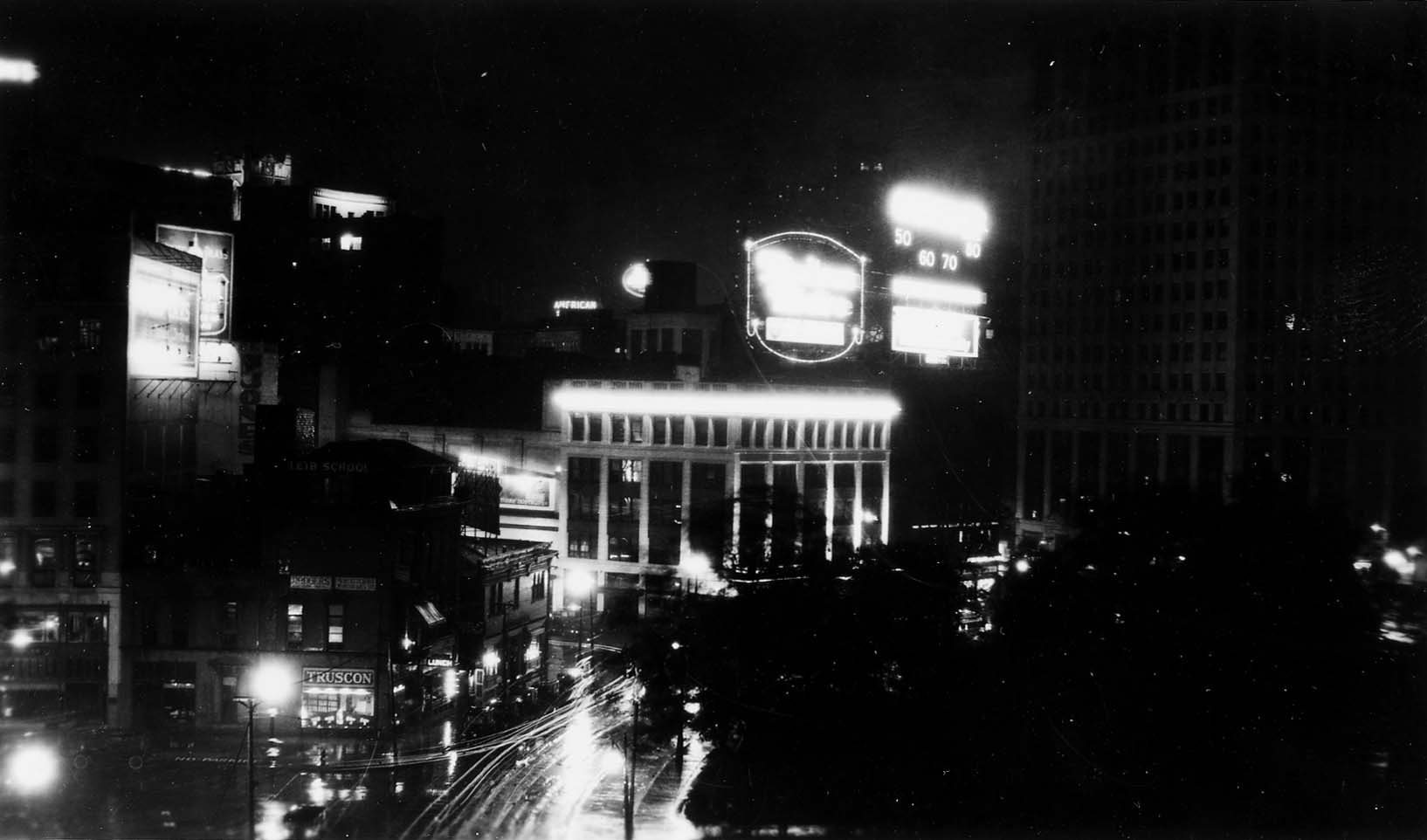 A wide night cityscape overlooking Grand Circus Park, showing the illuminated cornice of the Madison Theatre block, the Truscon building, and streaking traffic trails on wet streets.