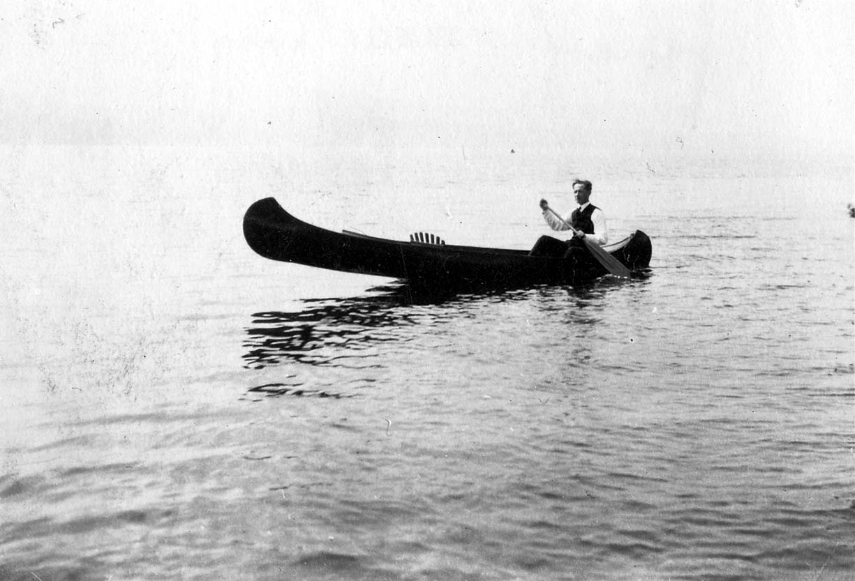 Donald A. Hall Sr. sitting alone in a dark canoe on a calm body of water, holding a paddle and looking toward the camera.