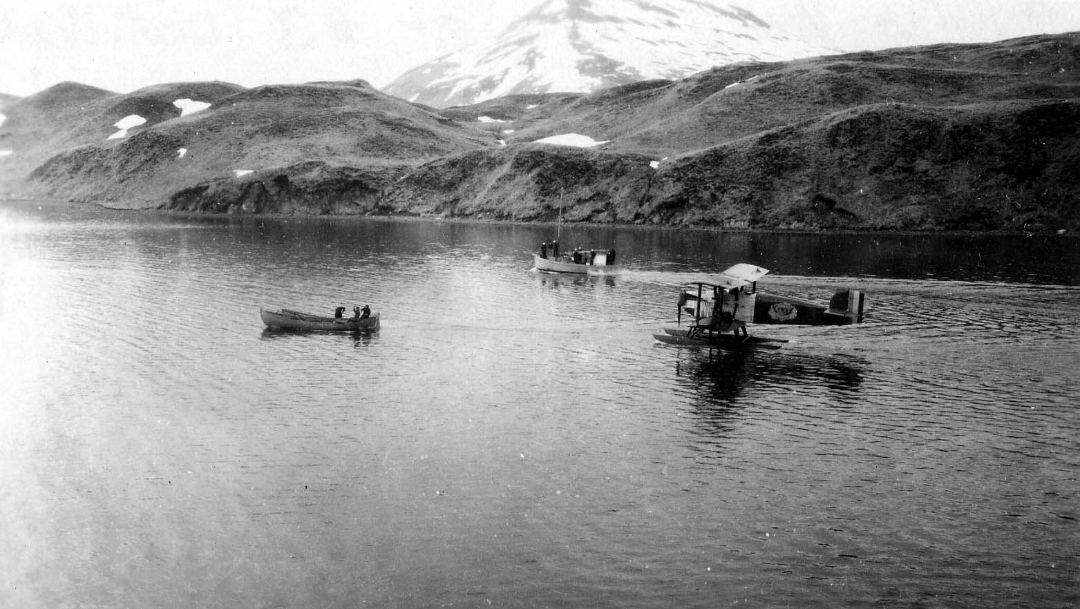 A Douglas World Cruiser biplane on pontoons floating in a calm Alaskan bay with snow-covered volcanic mountains in the background.