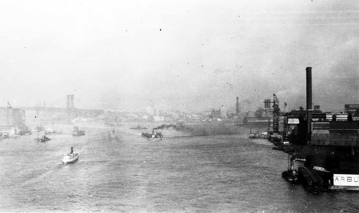 An elevated view looking up the East River from the Brooklyn Bridge, showing working steam tugs and the historic Arbuckle Brothers industrial complex along the DUMBO waterfront.