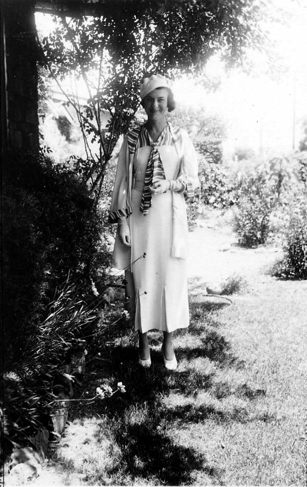 A full-length portrait of Elisabeth "Betty" Hall standing in a sunny garden, wearing a light-colored dress and a dark coat, smiling warmly.