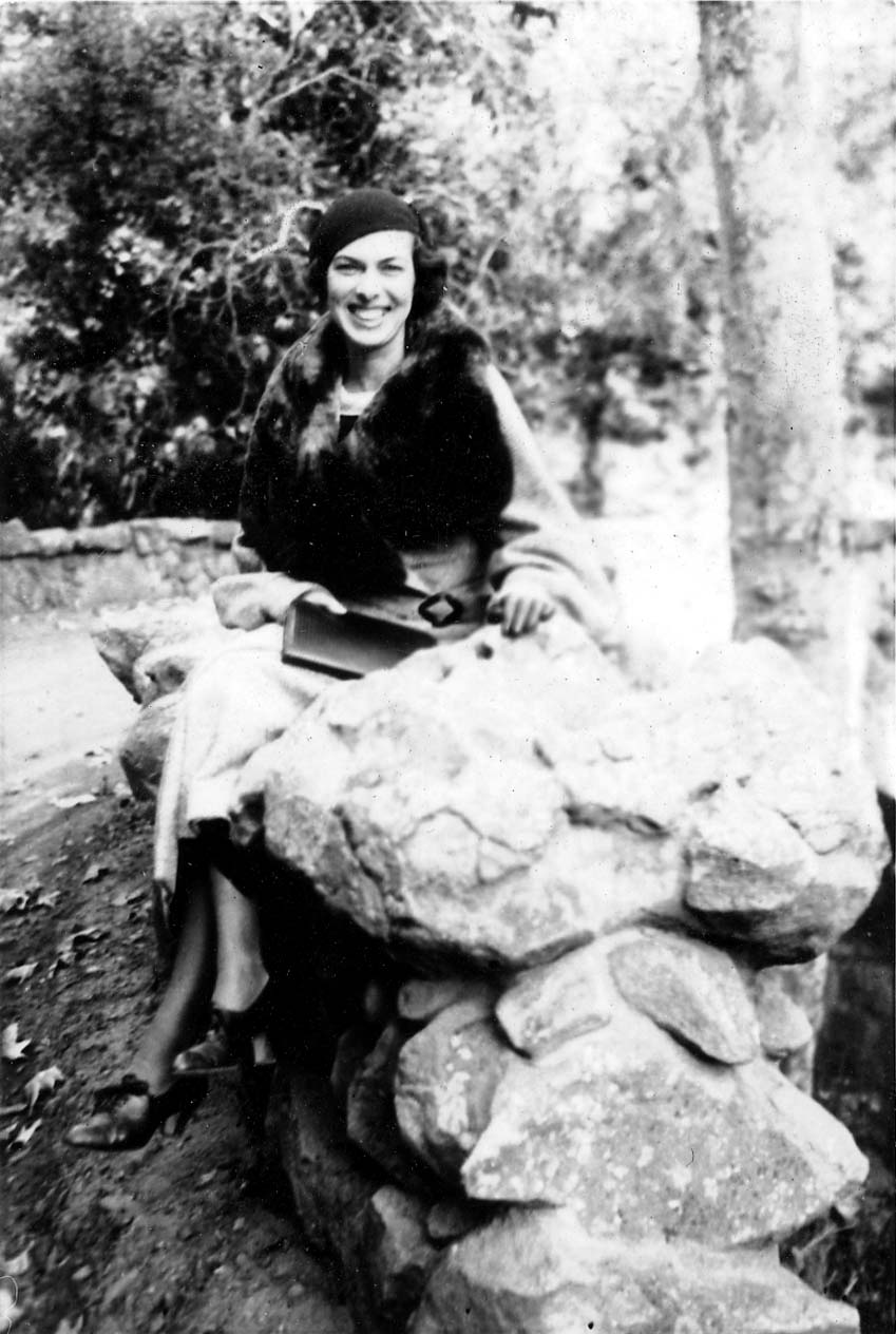 A portrait of Elisabeth "Betty" Hall smiling warmly, wearing a cloche hat and a fur-trimmed coat, sitting on a rustic stone wall.